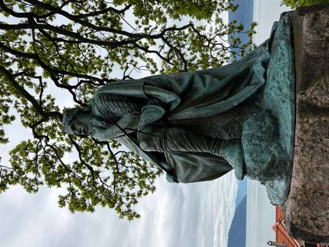 Bronze Viking statue sits pensively beneath leafy branches overlooking a fjord.