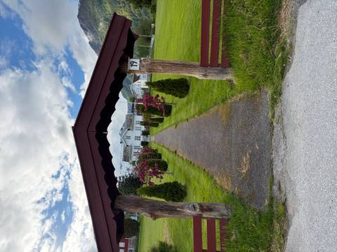 Pathway lined with young conifers leads to a white farmhouse framed by rustic timber archway and mountain peaks.