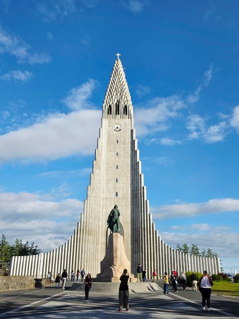 Tall concrete tower of Hallgrimskirkja church with a clock and a bronze statue at its base set against a bright blue sky with scattered clouds.