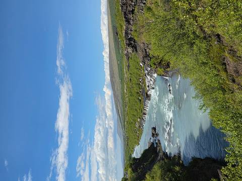 Wide view of a turquoise river cascading over small rocky falls through lush green vegetation under a bright blue sky.