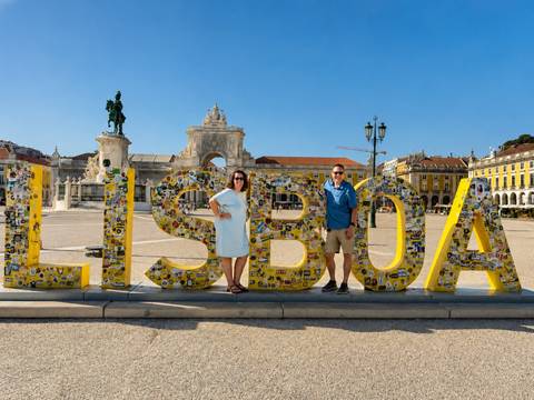 Two tourists pose in front of the large yellow 'LISBOA' sign at Lisbon's Praça do Comércio on a bright sunny day.