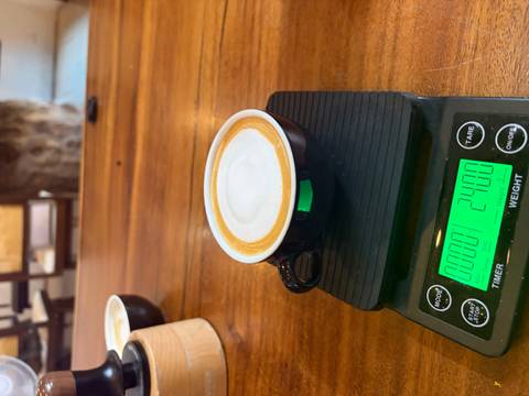 Fresh cappuccino in black cup placed on a digital coffee scale at a wooden counter.