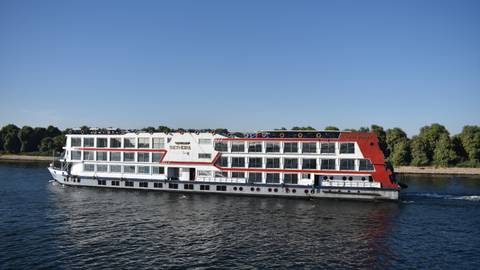 White-and-red Nile cruise ship sailing along calm river against clear blue sky.