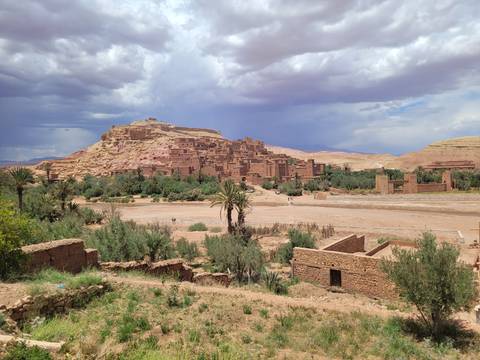 Panoramic view of the fortified clay village of Ait Benhaddou surrounded by palm trees and desert hills under a dramatic sky.