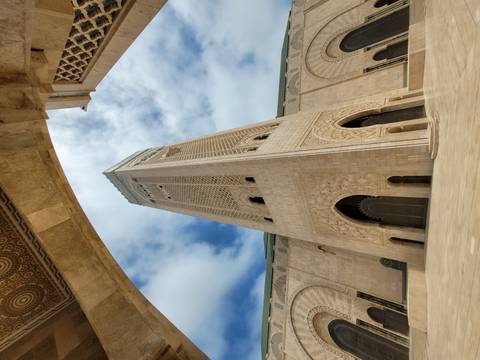 Upward perspective of the ornate minaret and archways of Hassan II Mosque against a partly cloudy blue sky.