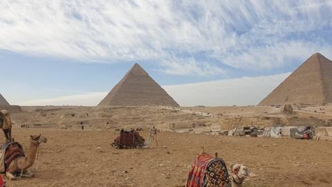 Iconic pyramids of Giza rising from sandy desert with camels and blankets in the foreground under streaked sky.