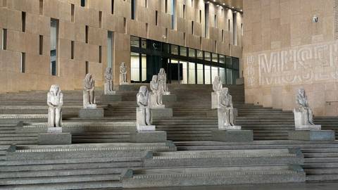 Series of seated ancient statues lining modern stone steps outside a contemporary museum building.