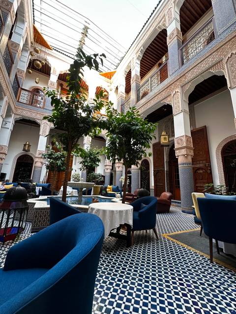 Elegant riad courtyard with tiled floor, central fountain, lush orange trees and patterned archways lit by a skylight.