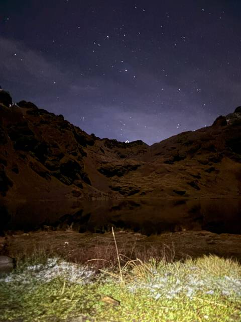 Nighttime scene of a star-studded sky over dark mountain silhouettes reflected in a still alpine lake.
