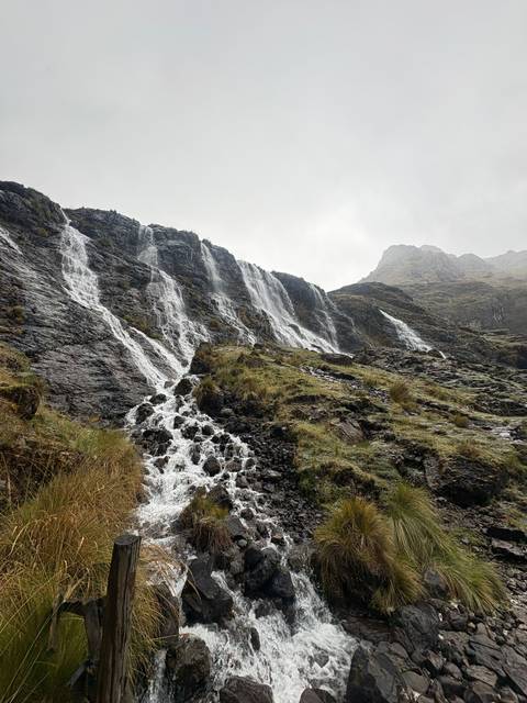 Multiple streams of water tumble down a steep, rocky Andean cliff under misty grey skies.