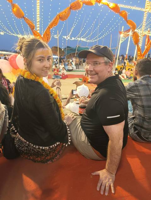 Two travelers wearing marigold garlands look back while seated among a festive crowd during an Indian ceremony.