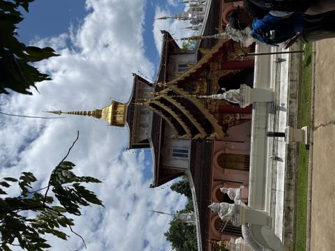 An ornate Thai temple with layered golden roof tiers and a shining spire rises against a partly cloudy sky.