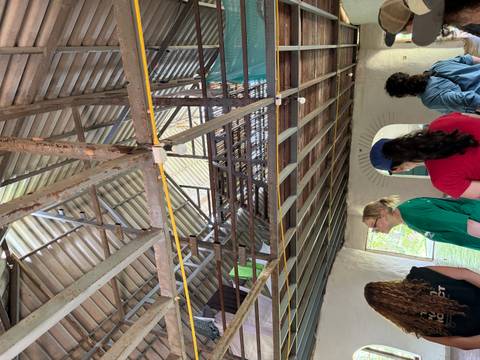 A group of visitors listen and look upward inside a rustic building with exposed beams and metal roof.