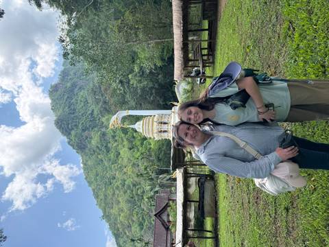 Two women stand smiling in a green valley with a white stupa and forested mountain rising behind.