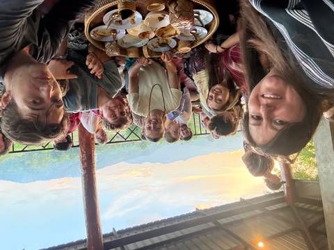 A cheerful group of travellers share dinner on a terrace overlooking sunset-lit mountains and valleys.