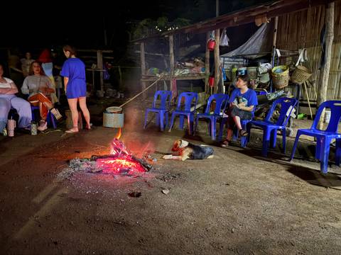 Travellers gather around a glowing campfire at night while a dog lies nearby and blue chairs ring the space.