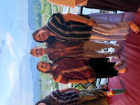Three women dressed in woven tops smile together on a patio with rice fields and mountains behind.