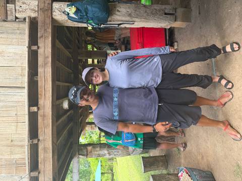 A traveller and a local guide stand arm in arm outside a rustic wooden building.
