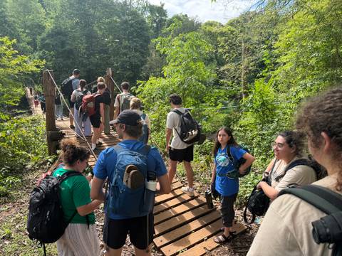 A line of hikers cross a wooden suspension bridge into dense tropical forest.