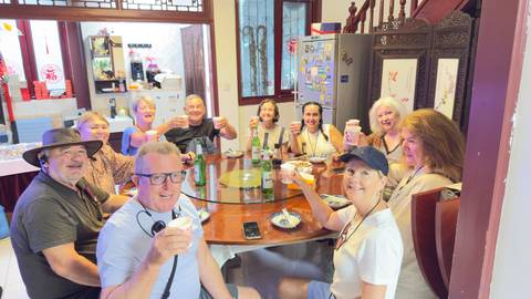 Group of travellers raise drinks around a circular dining table inside a traditional house.