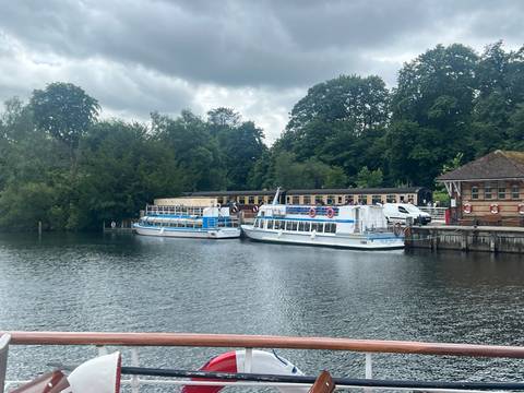 Docked sightseeing boats rest on calm lake waters bordered by leafy trees under grey skies.