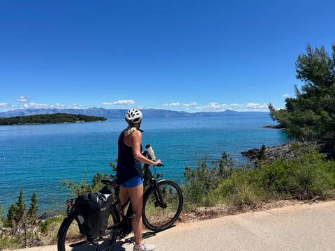 A female cyclist wearing a helmet pauses on a coastal dirt path, taking in the vivid turquoise sea, distant islands and rugged mountains under a clear blue sky.