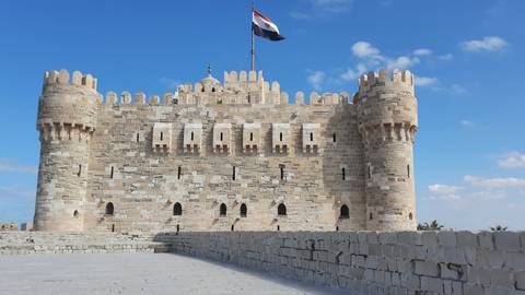 Stone walls and crenellations of the Citadel of Qaitbay fly the Egyptian flag against a vivid sky.