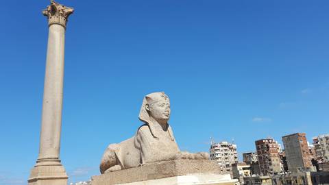 Granite sphinx statue beside Pompey’s Pillar with city apartment blocks under a bright blue sky.