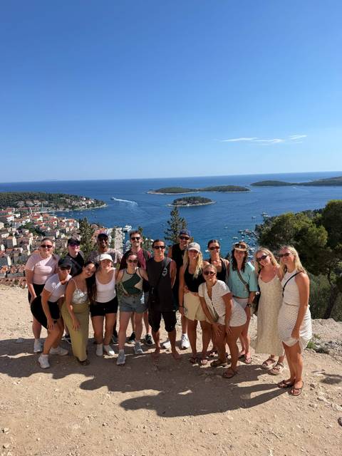 Group of travelers smiling at a cliff-top viewpoint overlooking Hvar town, small islands and deep blue Adriatic waters under a clear sky.