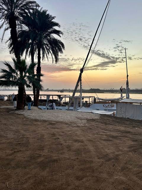 Traditional Nile sailboat moored at sandy riverbank at dawn, palms silhouetted against colorful sky.