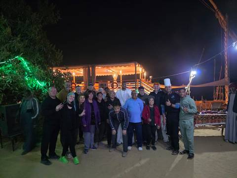 Large tour group poses at night on riverbank beside illuminated boat decorated with lights.