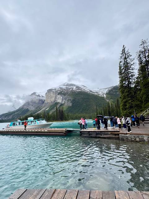 Tour group on a wooden dock boarding a small cruise boat on turquoise glacial lake framed by snow-capped cliffs.