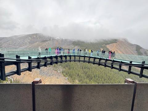Glass skywalk spanning a canyon with visitors in colorful ponchos against misty mountain backdrop.