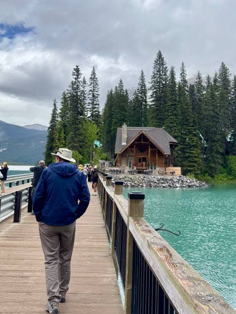 Visitors walking a wooden bridge toward a rustic lakeside lodge surrounded by dense evergreens and turquoise water.