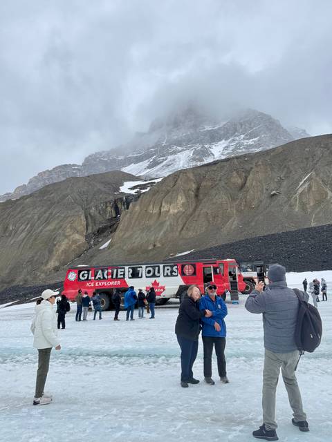 Large red Ice Explorer bus labeled 'Glacier Tours' parked on a snowy moraine with tourists milling about beneath towering cliffs.
