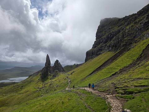 Hikers follow a stony trail toward the dramatic pinnacles of the Old Man of Storr beneath a stormy sky.