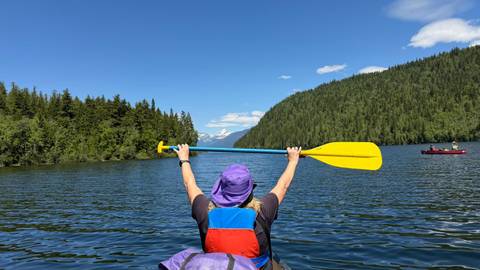 Kayaker raising a yellow paddle triumphantly while floating on a calm forested lake with snow-capped peaks in the distance.