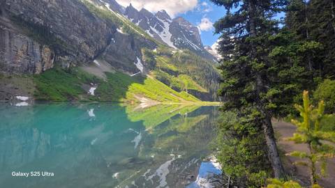 Crystal clear turquoise lake perfectly reflecting alpine peaks and greenery; small white text in the corner notes phone model.