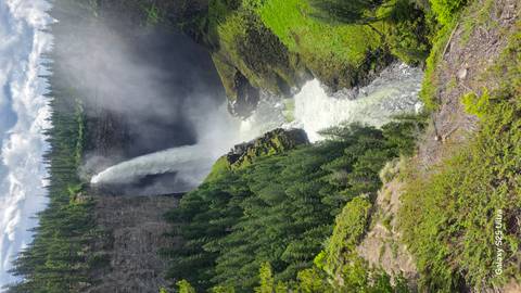 Tall powerful waterfall plunging into a deep canyon surrounded by dense evergreen forest; phone watermark visible in corner.