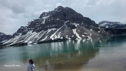 Snow-streaked mountain rising above a cold turquoise lake with lone person near shore; phone watermark text on lower left.
