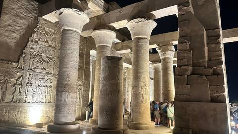 Massive stone columns with carved capitals inside Edfu temple lit at night while visitors look up.