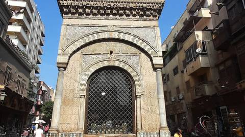Ornate Mamluk sabil-kuttab with intricate stonework and iron grille set amid Cairo street buildings.