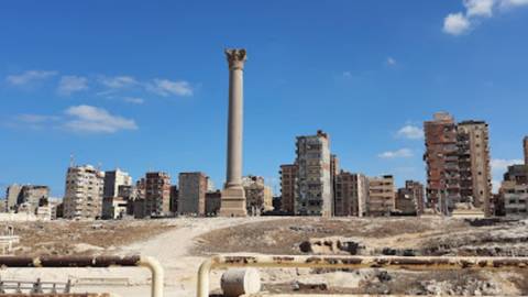 Tall solitary column (Pompey’s Pillar) rising above rubble with apartment blocks behind