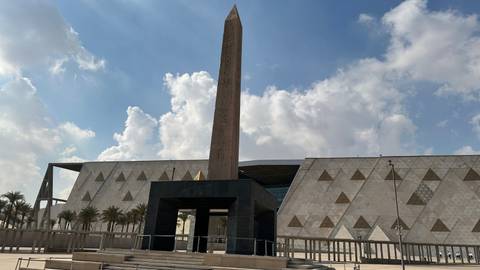 Modern museum facade with central obelisk framed by patterned stone walls and palm trees