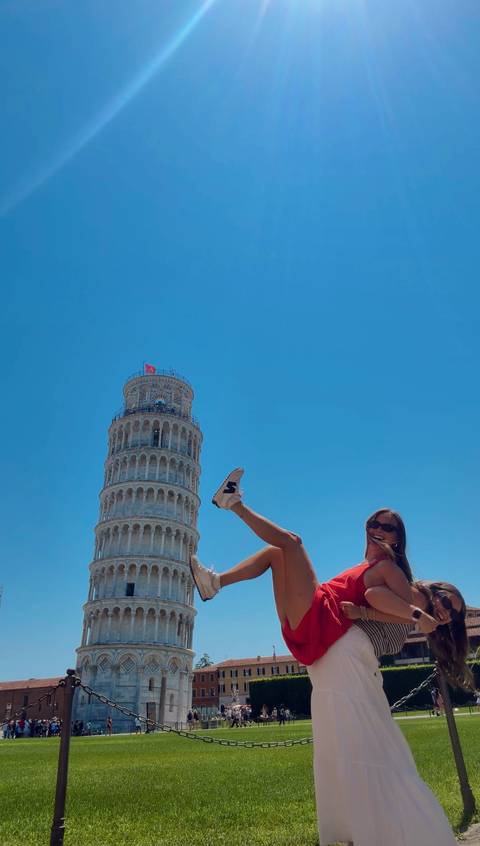 Smiling traveller playfully kicking a leg up with the Leaning Tower of Pisa rising against a cloudless blue sky.