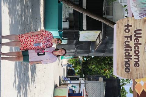 Two visitors pose beneath a large sign reading 'Welcome to Fulidhoo' on a sandy village street.