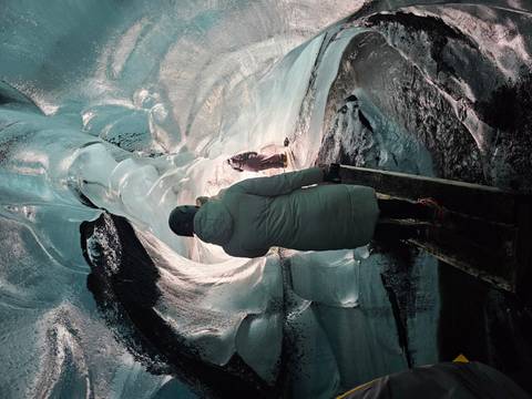 Traveler in parka and helmet walks a wooden plank through a turquoise ice tunnel