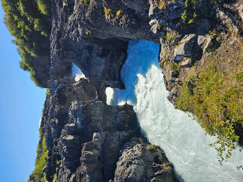 Blue glacial river rushes through jagged volcanic rock formations under bright sun