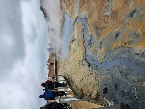 Visitors walk boardwalks around steaming, colorful geothermal pools and barren earth