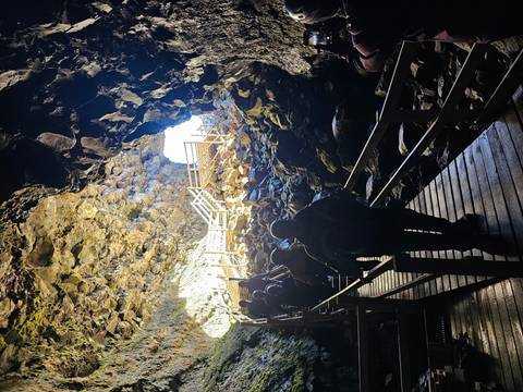 Travelers descend wooden stairs into a rocky lava cave illuminated by daylight at entrance